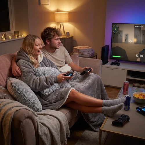 Couple relaxing on the couch for a game night, wrapped in a grey Toasty Hoodie sherpa wearable blanket